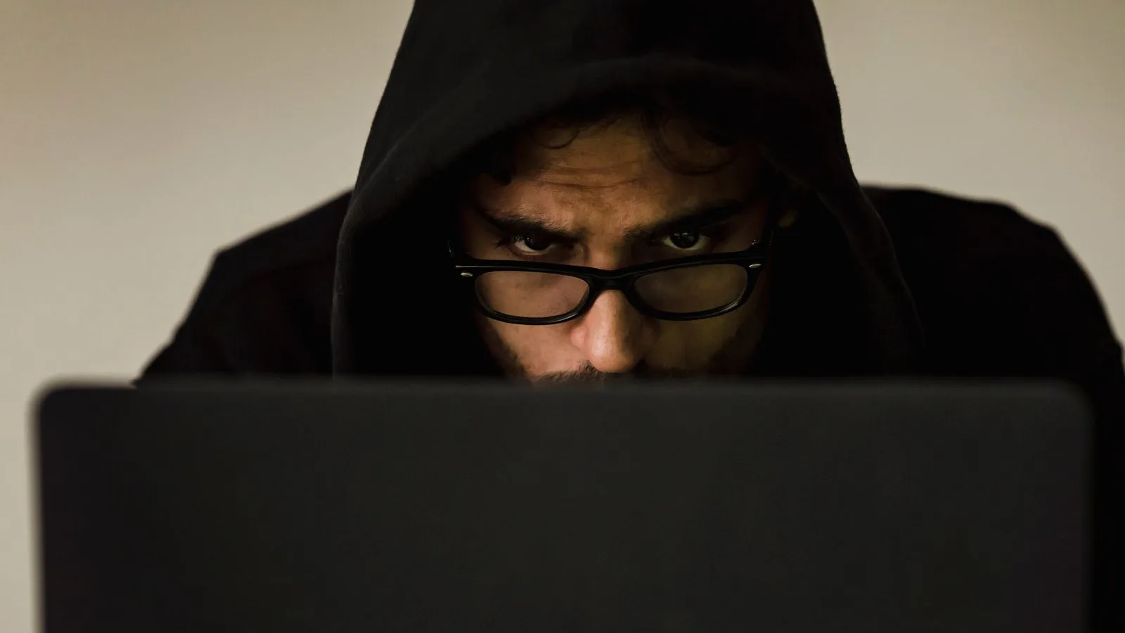 Persoană cu glugă și ochelari, aplecată asupra unui laptop într-o cameră întunecată