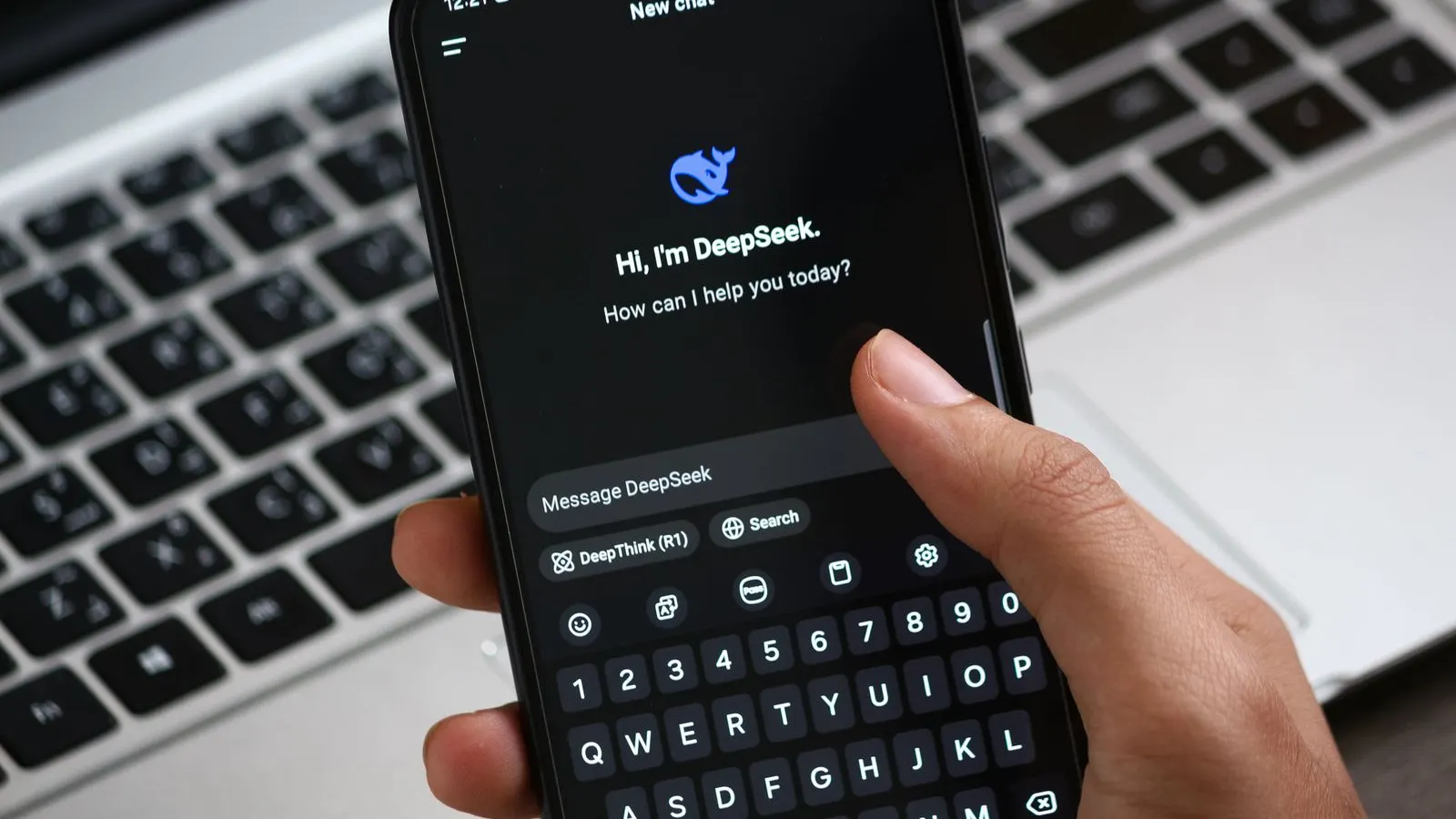 Mână care ține un telefon cu aplicația de chat DeepSeek deschisă, deasupra unui laptop