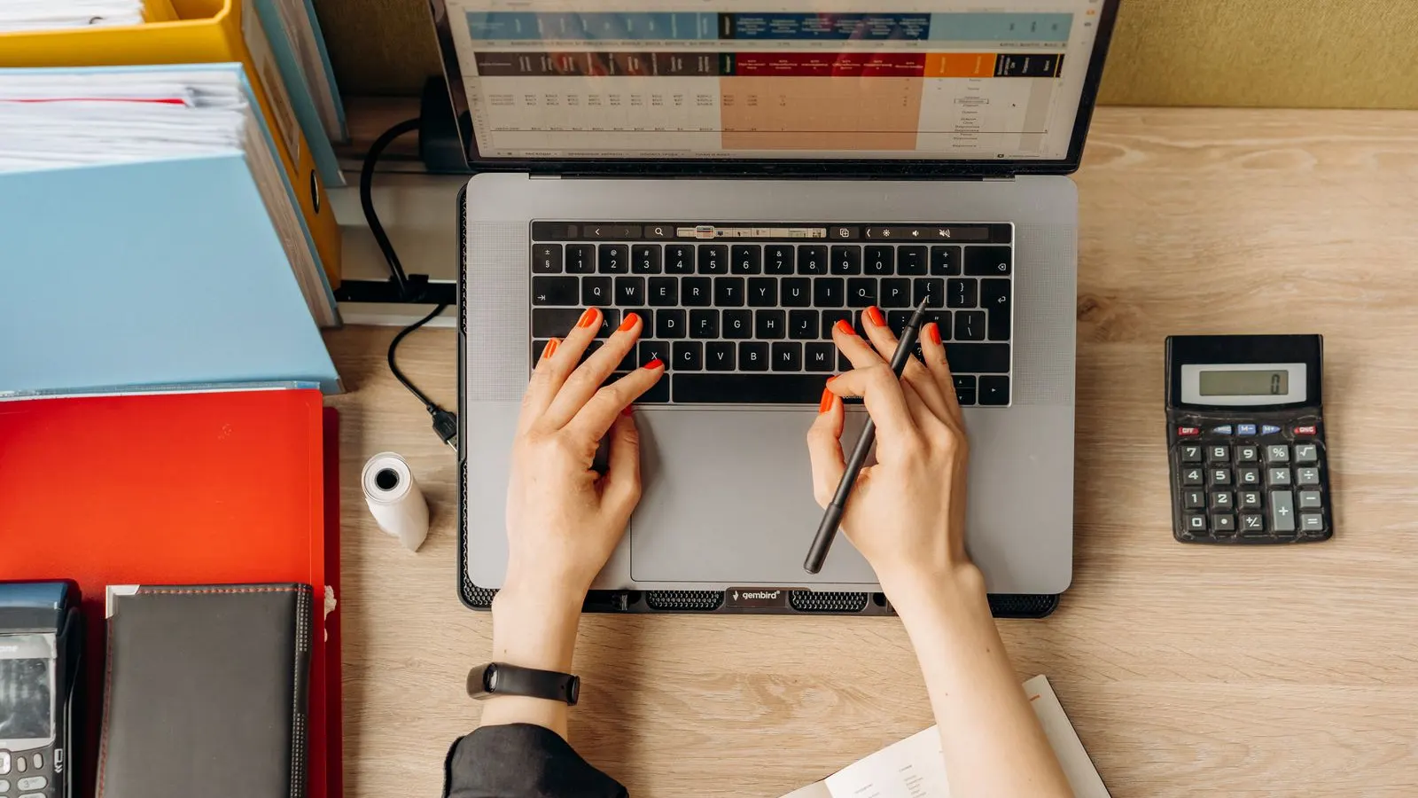 Mâini pe tastatura unui laptop cu tabel contabil, lângă un calculator de birou