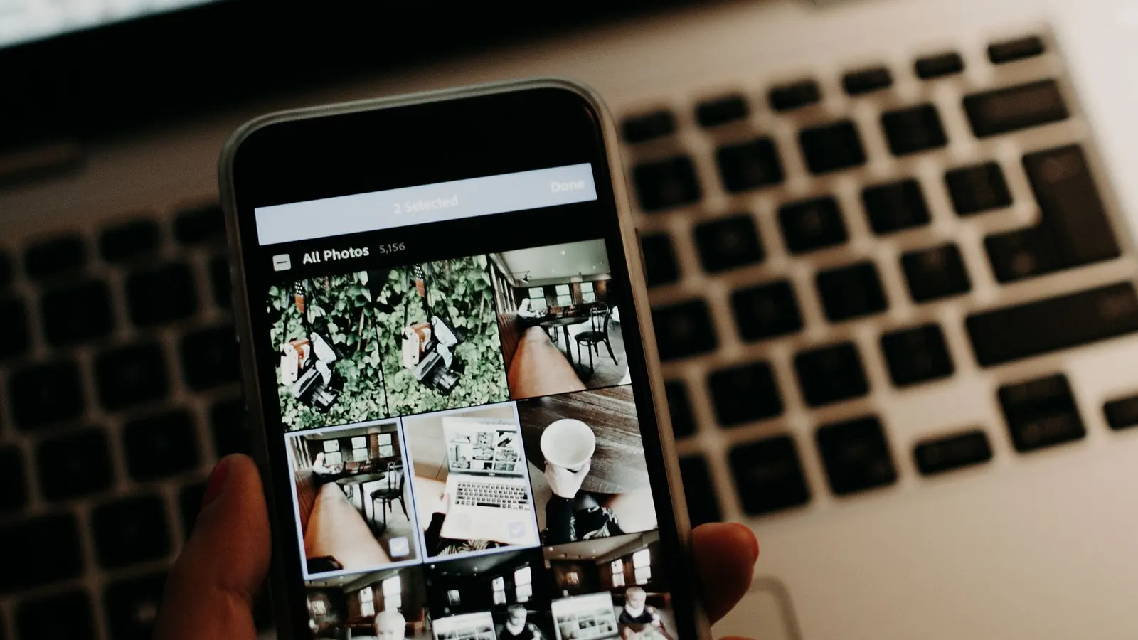 Mână care ține un telefon cu galeria de fotografii deschisă, în fața unui laptop