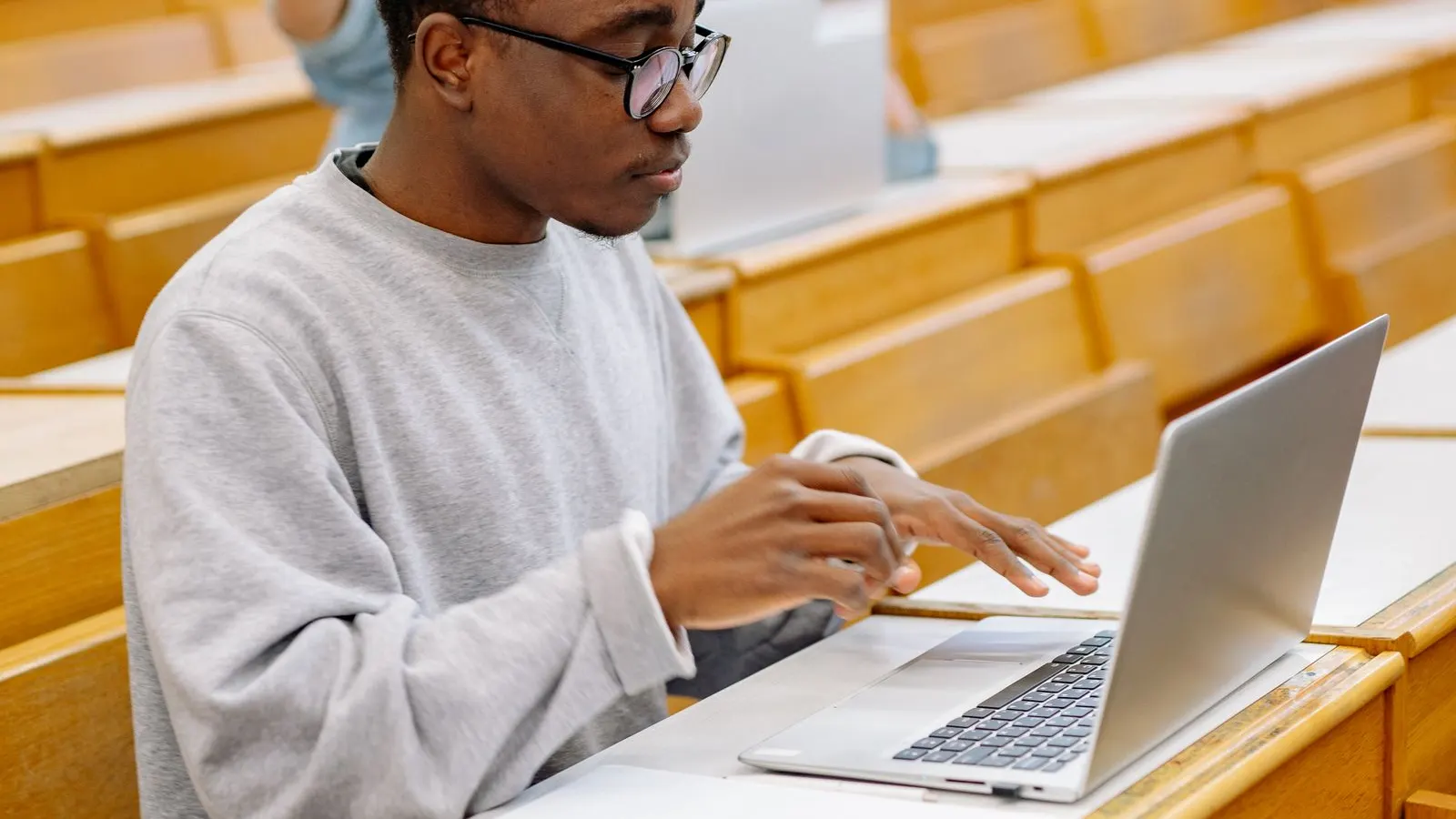Student cu ochelari care scrie la un laptop argintiu într-o sală de curs