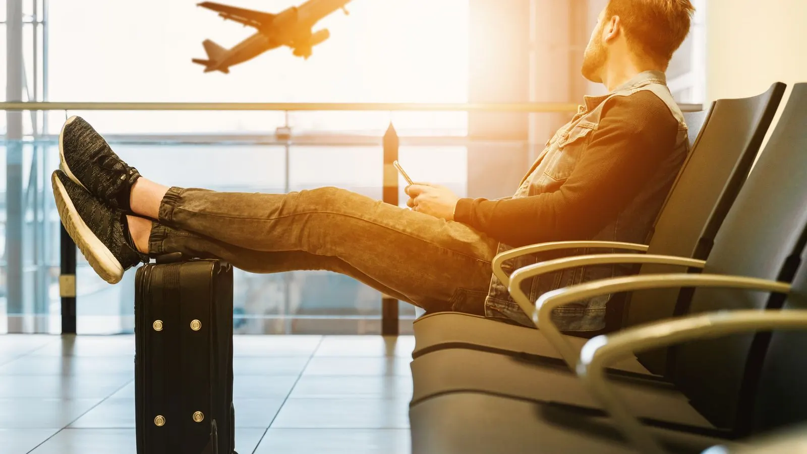 Călător cu telefonul în mână așteaptă în aeroport, iar un avion decolează în fundal